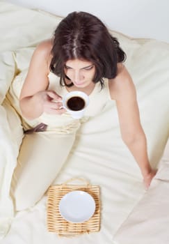 High angle view at young brunette woman in bed with cup of coffee