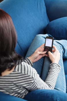Young brunette woman sitting on blue sofa and texting with her cellphone