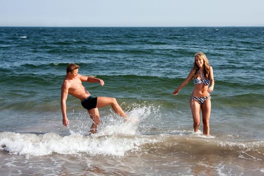 Beautiful young couple playing in the ocean splashing water, heaving fun on a summer day