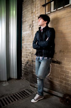 Full body street shot of young man leaning against brick wall looking away.