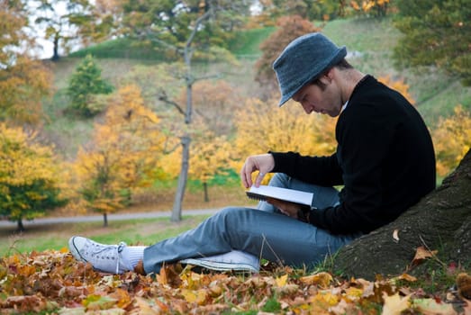 Young man reading under a tree in the park during autumn.