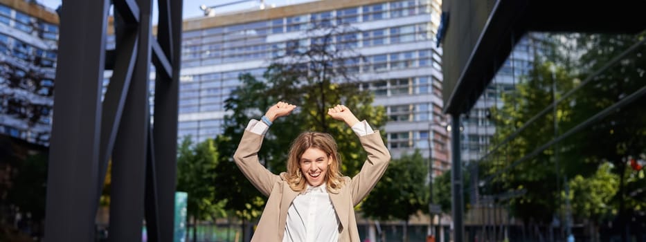 Portrait of happy young businesswoman lift hands up, does fist pump, celebrates victory, wins smth, achieves goal, stands outdoors on street.