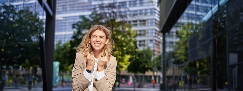 Excited businesswoman, office lady in suit, rejoicing, standing on street and celebrating, feeling happy, posing outdoors near office buildings.