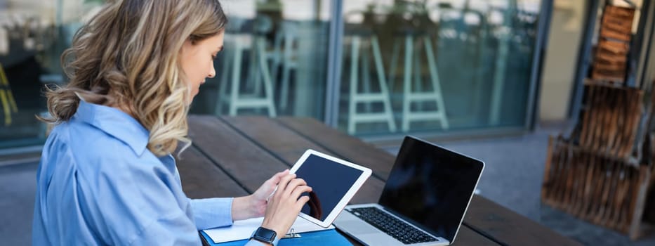 Portrait of businesswoman working on digital tablet, checking diagrams, sitting outdoors on fresh air near office building. Corporate woman prepare for work meeting.