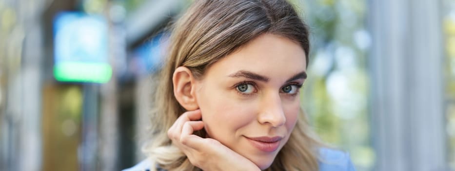 Close up portrait of young woman, tuck hair behind ear, looking flirty and smiling, sitting in blue shirt outdoors on street.