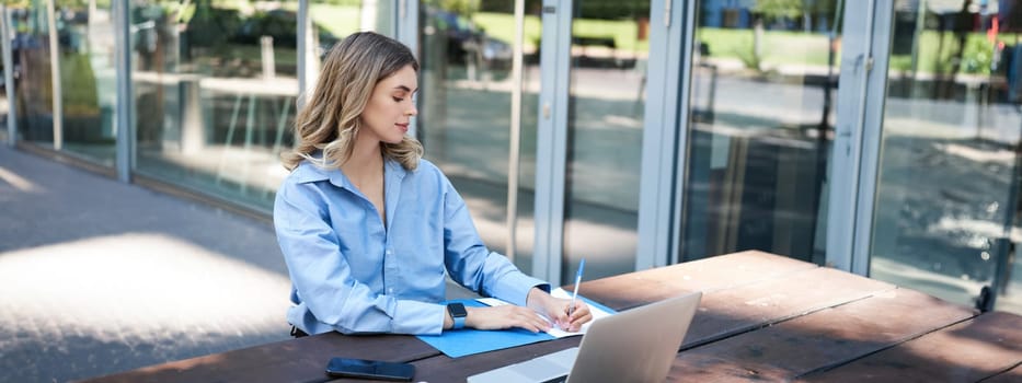 Portrait of beautiful young woman working on fresh air outside. Corporate woman writing in her documents and folders, using laptop outdoors.