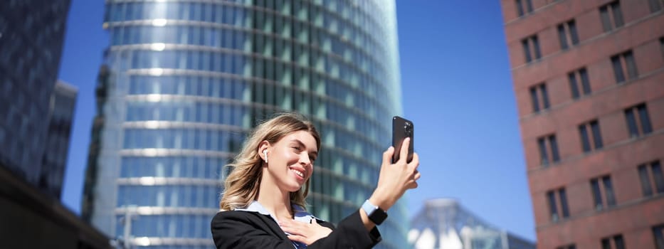 Portrait of businesswoman wave her hand at mobile phone camera, waves hand during video chat, stands in suit in city center outdoors.