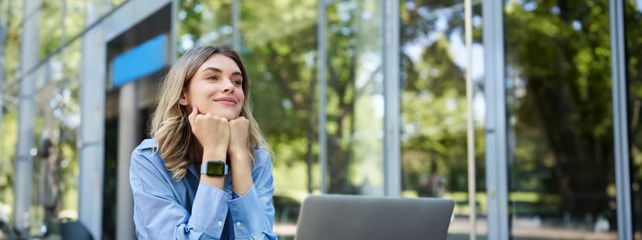 Dreamy woman sitting with laptop outdoors on street. Smiling businesswoman daydreaming, imaging smth while working outside office building.