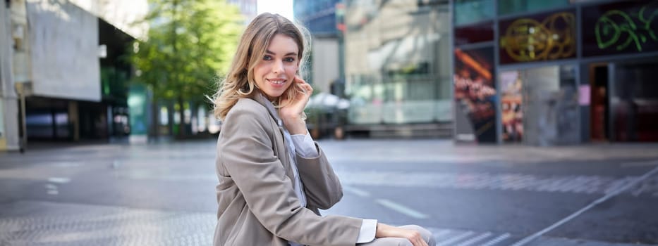 Confident blond businesswoman in suit, smiles at camera, sits outdoors near office buildings.