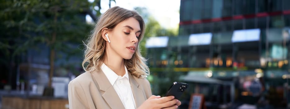 Successful businesswoman in beige suit, wireless headphones, looking at mobile phone, using smartphone app, standing outdoors on street.