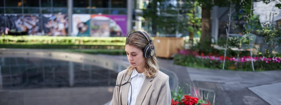 Portrait of businesswoman being on a meeting online outside office, listening in headphones, looking at laptop, working outdoors.