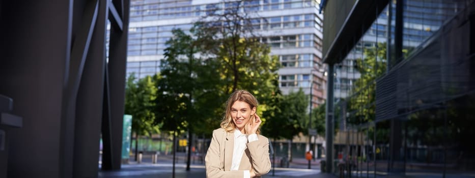 Portrait of smiling businesswoman in corporate clothing, looking confident and happy, wearing beige suit, standing outdoors on street, outside office.