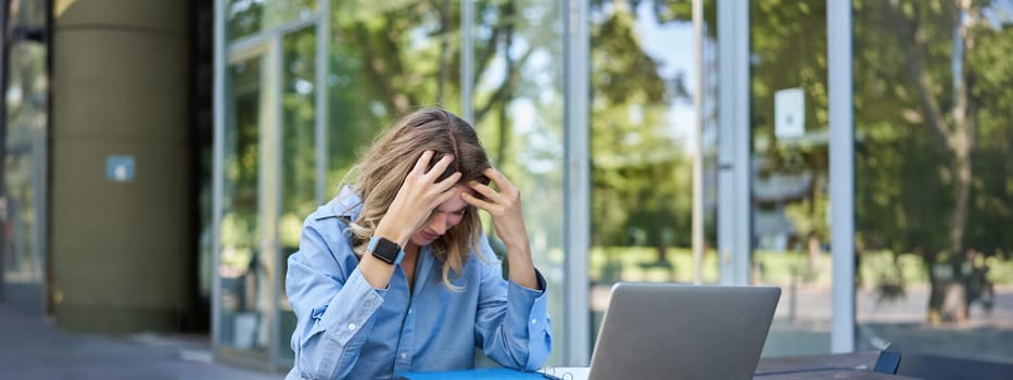 Portrait of young working woman sitting near laptop and feeling frustrated, disappointed. Businesswoman looks troubled while works on project, sits outdoors.