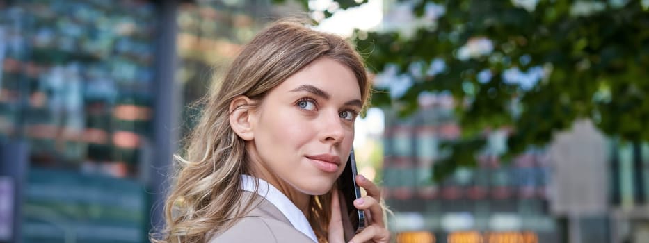 Close up shot of businesswoman talking on mobile phone. Corporate woman calling someone, looking around, standing outdoors.