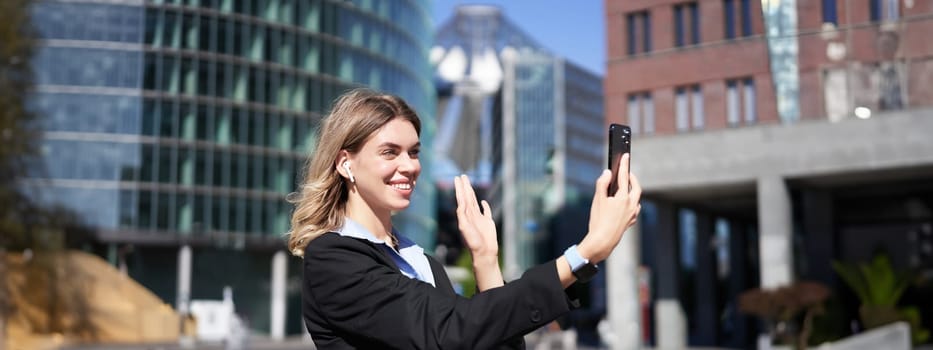 Portrait of businesswoman wave her hand at mobile phone camera, waves hand during video chat, stands in suit in city center outdoors.