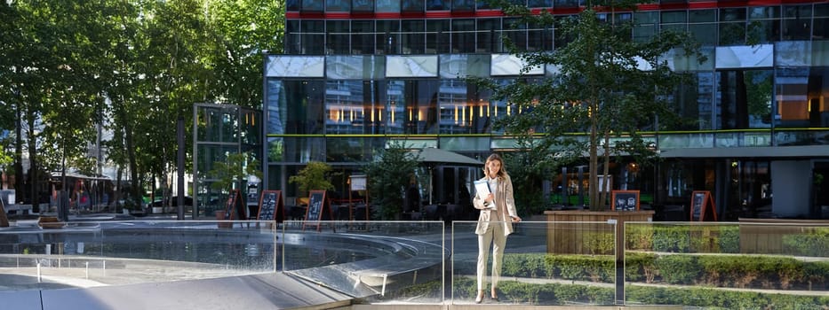 Corporate businesswoman in beige suit. Silhouette of young corporate woman with documents and laptop, posing outdoors in city center.