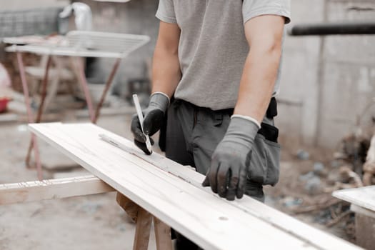 One young unrecognizable Caucasian man in gray gloves holds a construction measuring tape and draws on a board with a pencil, standing in the backyard of a house on a summer day, side view close-up.