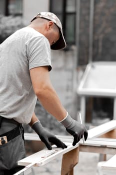 One young Caucasian man in a uniform and gray gloves, standing from behind holding a construction measuring tape and drawing on a board with a pencil, standing in the backyard of a house on a summer day, side view close-up.