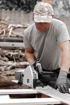 One young Caucasian man in a uniform and gray gloves cuts a board with an electric saw while standing in the backyard of a house on a summer day, side view close-up.