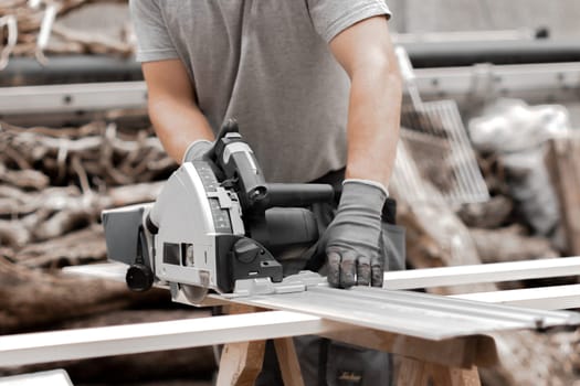 One young Caucasian unrecognizable man in a uniform and gray gloves is sawing a board with an electric saw while standing in the backyard of a house on a summer day, close-up side view.