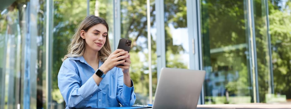 Portrait of saleswoman working outside office, sititng with laptop, using mobile phone. Digital nomad woman studying, using computer outdoors.