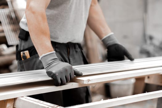 One young Caucasian unrecognizable man in a uniform and gray gloves presses a board with a metal ruler for cutting while standing in the backyard of a house on a summer day, close-up side view.