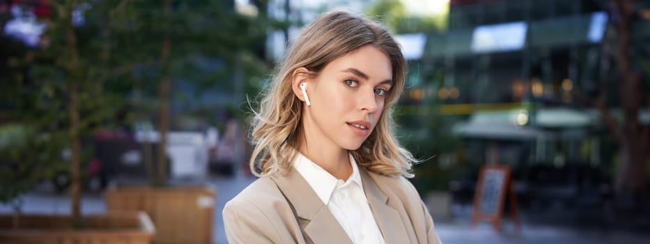 Confident businesswoman in headphones and beige suit, looking at camera with thoughtful gaze, posing outdoors.