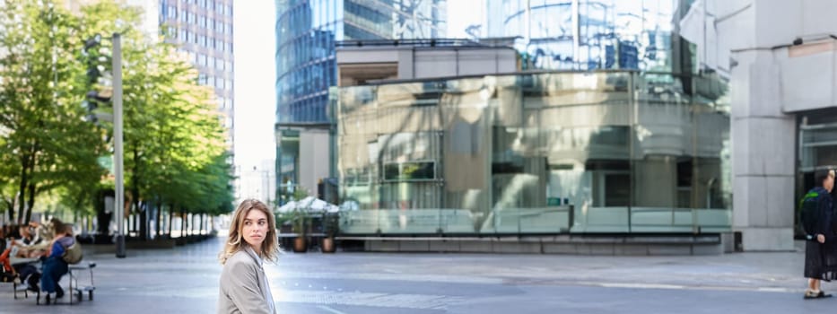 Female corporate worker sitting outdoors near office buildings, turn back and look behind, waiting for coworker on lunch break.