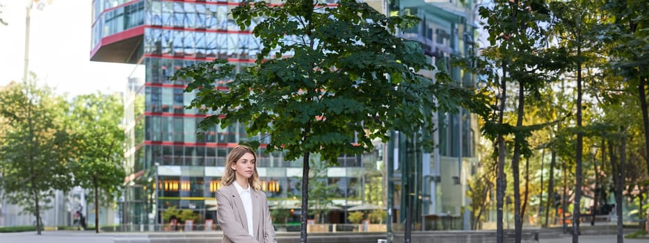 Portrait of young saleswoman in beige business suit, holding blue folder with work documents, standing outdoors on street of city center.