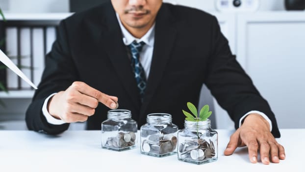 Businessman put coin to money saving glass jar at his office as sustainable money growth investment or eco-subsidize. Green corporate promot and invest in environmental awareness. Gyre