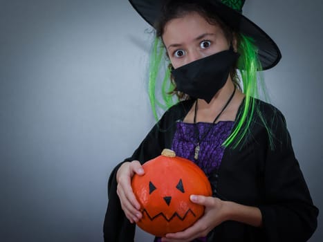 Portrait of a surprised caucasian girl with wide open eyes in a Halloween hat, a black mask holding a pumpkin with a painted muzzle in her hands stands on the right against the wall on a gray background with little copy space on the left, close up side view. Halloween concept, Halloween celebration.