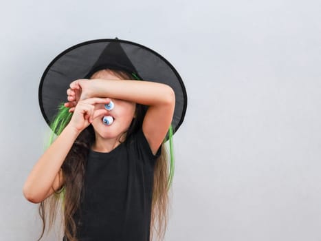 Portrait of a little caucasian girl in black clothes with a halloween hat holds a candy near her mouth with one hand, and closes her eyes with the other, stands on the left against the wall on a gray background with copy space on the right, close up side view. Halloween concept, Halloween celebration.