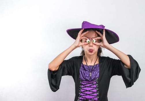 Portrait of a caucasian girl in black clothes with a halloween hat holds candy over her eyes with her hands and shows her tongue stands on the right against a white wall with copy space on the left, close up side view. Halloween concept, Halloween celebration.