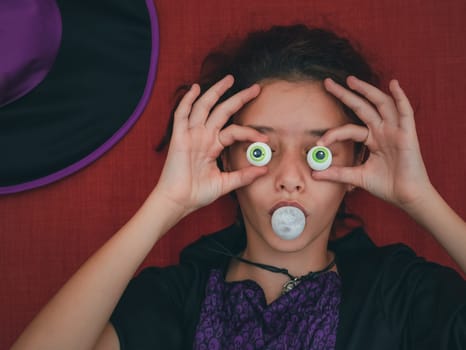 A teenage girl in a witch costume covers her eyes with candy and blows a ball of chewing gum lying on an orange background, close-up top view.