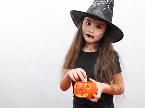 Portrait of a beautiful caucasian brunette girl in black clothes with long hair, halloween makeup and a witch hat holds a ceramic pumpkin with candies in her hands looking at the camera. stands on the right on a white background with copy space on the left, close-up side view. Halloween concept, Halloween celebration.