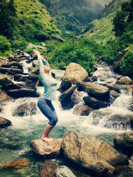 Vintage retro effect hipster style image of sporty fit woman doing yoga asana Utkatasana (chair pose) outdoors at tropical waterfall standing on stone