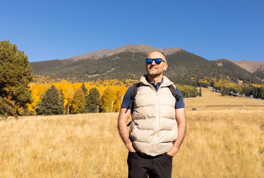 Handsome Smiling Man Enjoys Hiking In Mountains At Fall Season On Sunny Day, Yellow Forest on Background. Male Hiker with Backpack Does Trekking In Autumn Nature. Travel, Active Lifestyle Copy Space.