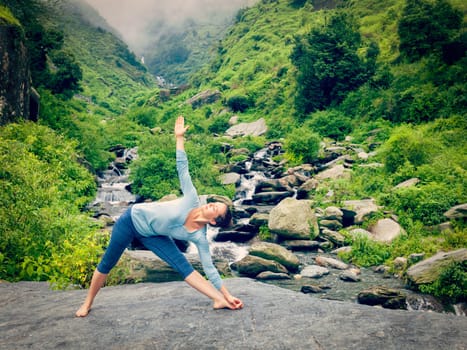 Vintage retro effect hipster style image of woman doing Ashtanga Vinyasa yoga asana Utthita trikonasana - extended triangle pose outdoors at waterfall in Himalayas