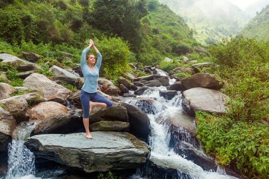 Woman in Hatha yoga balance yoga asana Vrikshasana tree pose at waterfall outdoors