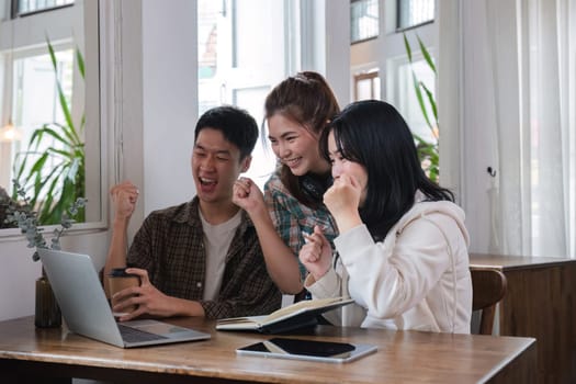 A group of cheerful young university students are looking at a laptop screen and showing their fists, celebrating a good news together while sitting in a cafe..