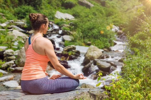 Woman in Hatha yoga asana Padmasana outdoors at tropical waterfall