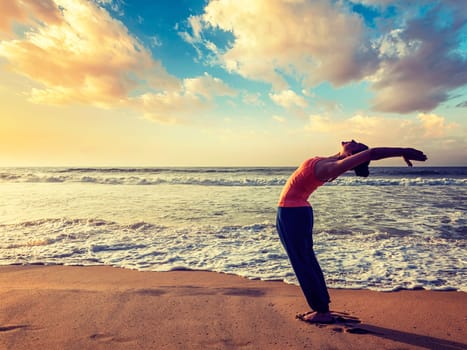 Vintage retro effect hipster style image of young sporty fit woman doing yoga Sun salutation Surya Namaskar pose Hasta Uttanasana on tropical beach on sunset