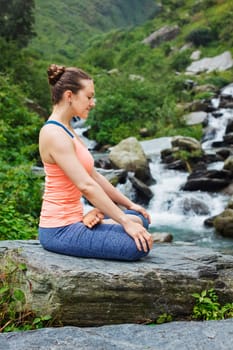 Woman in Hatha yoga asana Padmasana outdoors at tropical waterfall