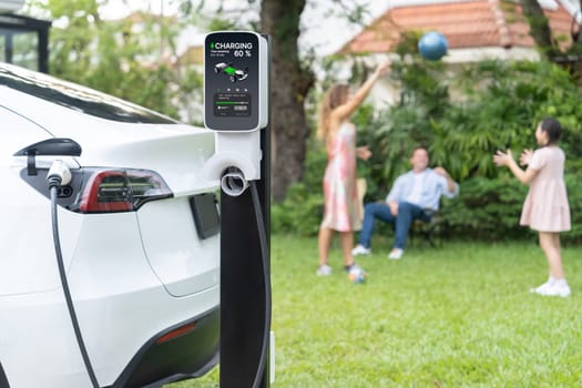 Focus electric vehicle recharge from home charging station on blur background of happy and playful family playing together. EV car using alternative and sustainable energy for better future.Synchronos