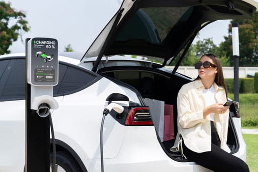 Young woman use smartphone to pay for electricity at public EV car charging station green city park. Modern environmental and sustainable urban lifestyle with EV vehicle. Expedient