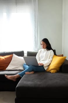 Young Asian woman working with laptop at home sitting on the couch in cozy apartment living room. Copy space. Vertical image.