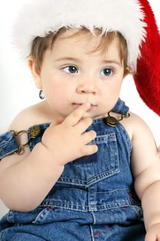 Baby girl in denim bib and brace overalls with finger to mouth thinking gesture.  White background.  Christmas theme.