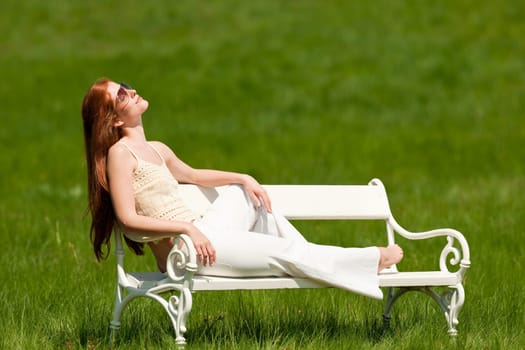 Red hair woman enjoying sun on white bench in green meadow, shallow DOF