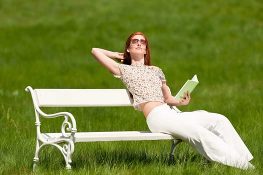 Red hair woman sitting with book on white bench in a meadow shallow DOF