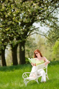 Red hair woman reading book on white bench in green meadow, shallow DOF